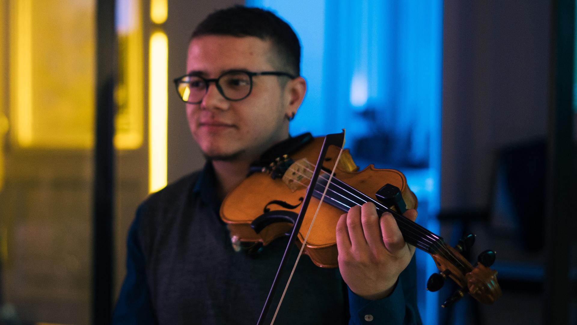 Ein junger Mann spielt Violine, während der Hintergrund des Raumes in den Nationalfarben der Ukraine, gelb und blau, ausgeleuchtete ist.