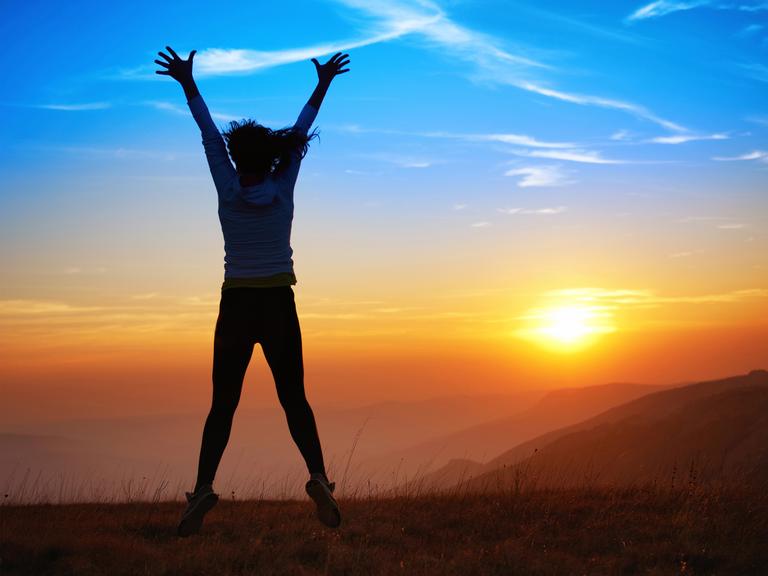 Rückansicht einer sportlichen Frau, die auf einem Berg steht und den Sonnaufgang mit hochgerissenen Armen enthusiastisch begrüßt.