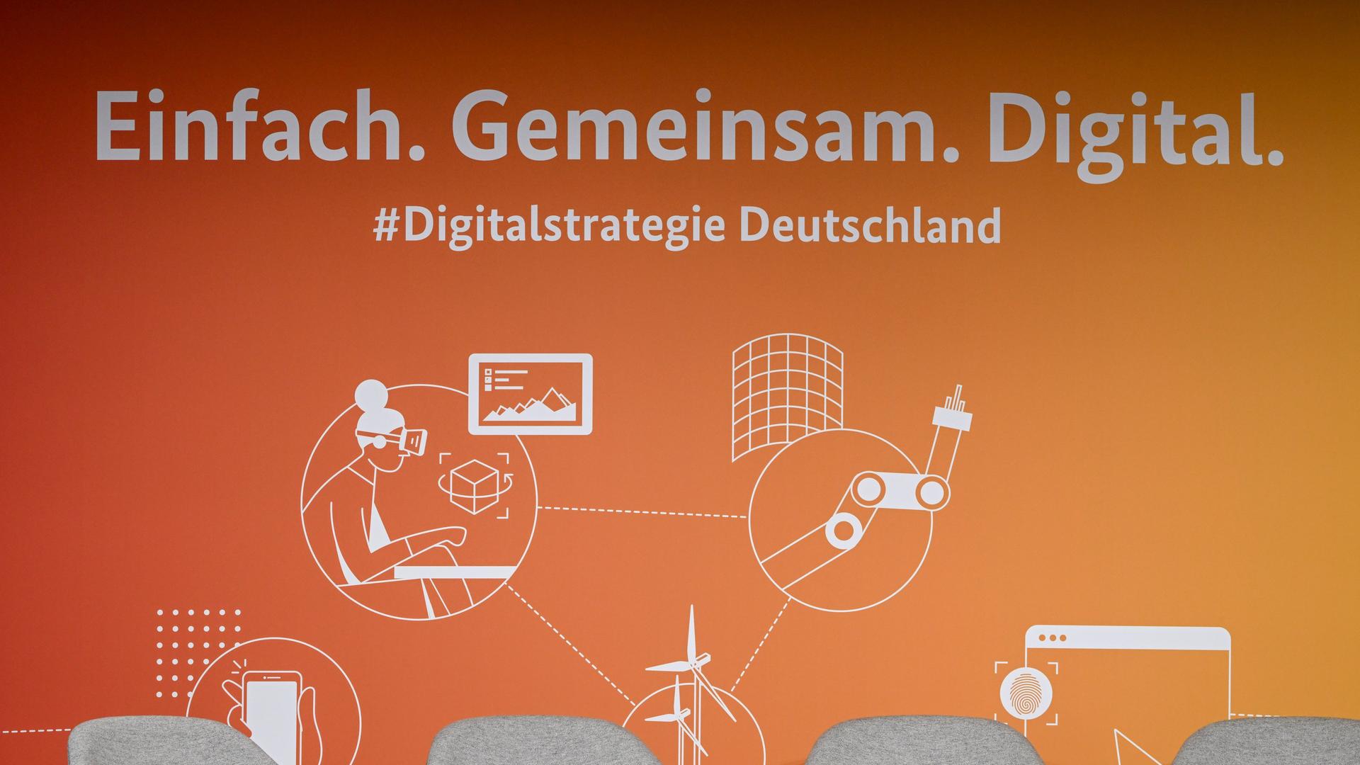 Logos für die Digitalstrategie Deutschland prangen an einer orangenen Wand. Im Vordergrund stehen leere graue Stühle. 