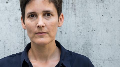 Eine Frau mit kurzem braunem Haar und braunen Augen blickt ernst in die Kamera. Sie trägt ein dunkelblaues Hemd mit Kragen. Im Hintergrund ist eine graue Wand zu sehen.