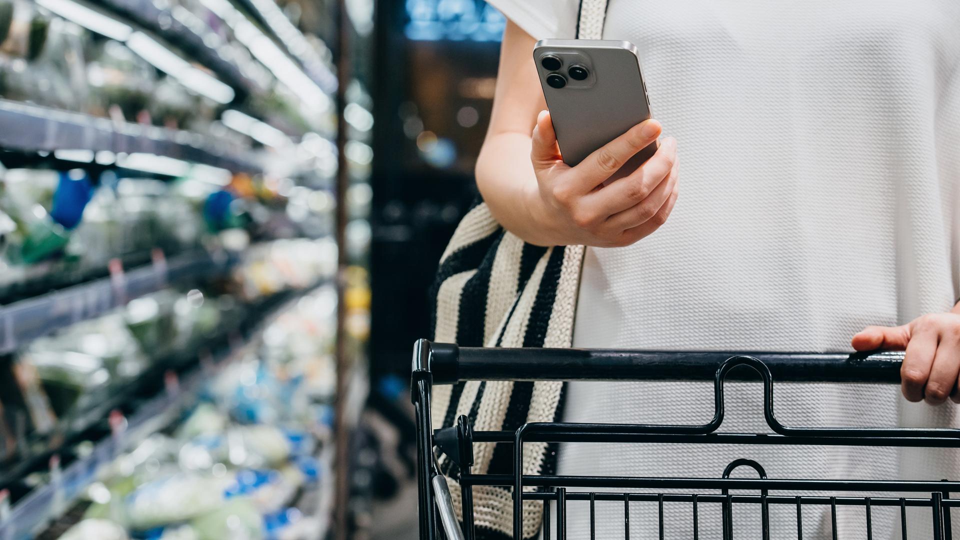 Eine Person mit Einkaufswagen beim Lebensmitteleinkauf im Supermarkt. In der Hand hält sie ein Smartphone.