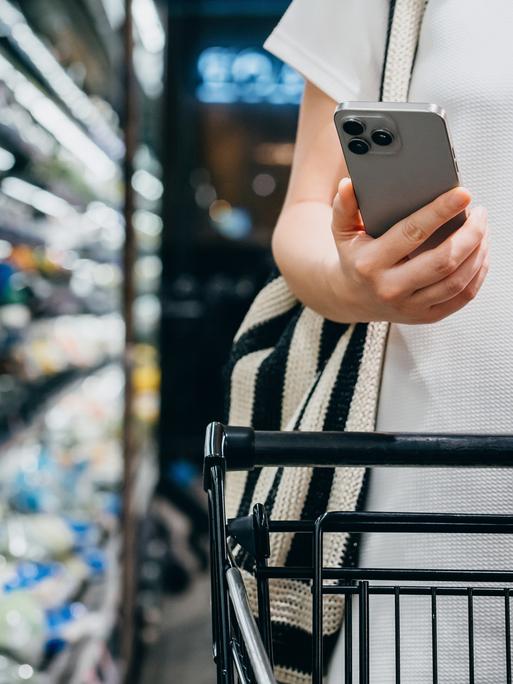 Eine Person mit Einkaufswagen beim Lebensmitteleinkauf im Supermarkt. In der Hand hält sie ein Smartphone.