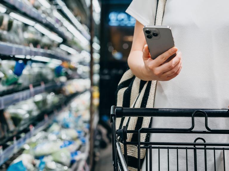 Eine Person mit Einkaufswagen beim Lebensmitteleinkauf im Supermarkt. In der Hand hält sie ein Smartphone.