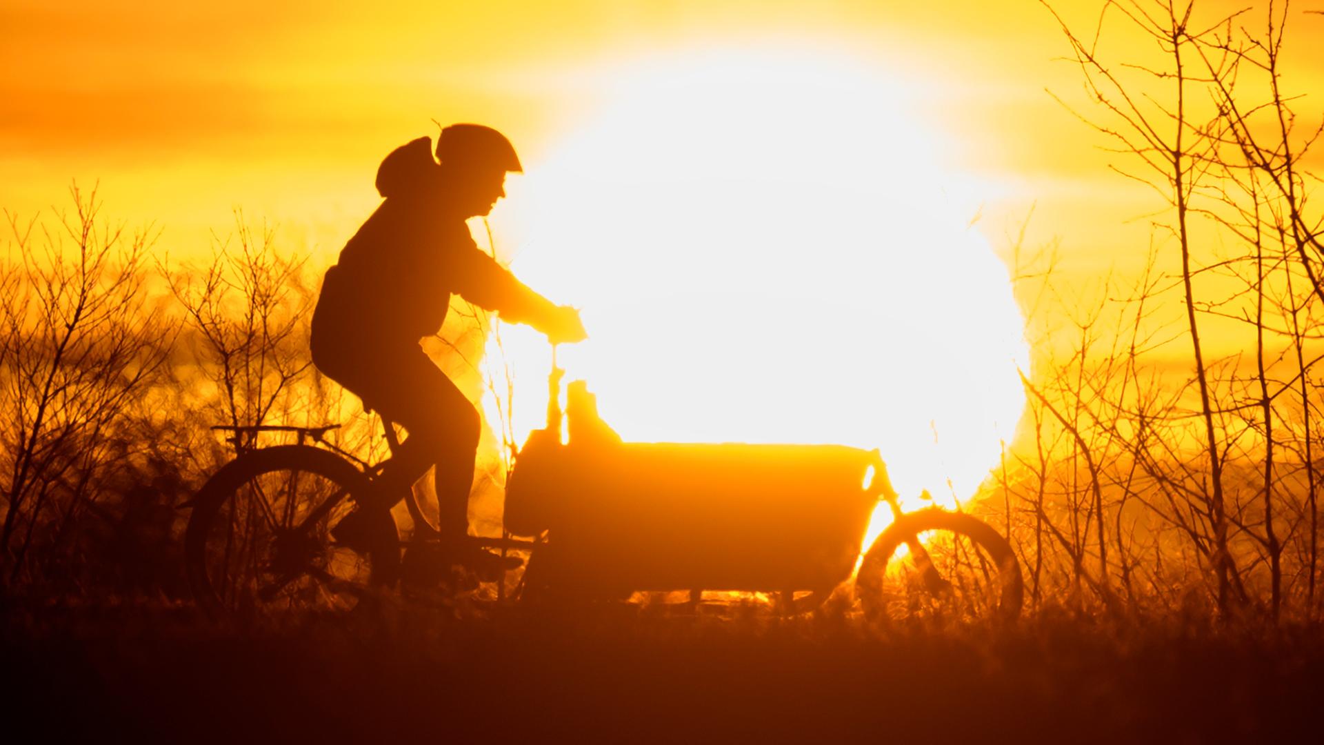 Ein Mann fährt bei Sonnenaufgang mit einem Lastenfahrrad über einen Feldweg.