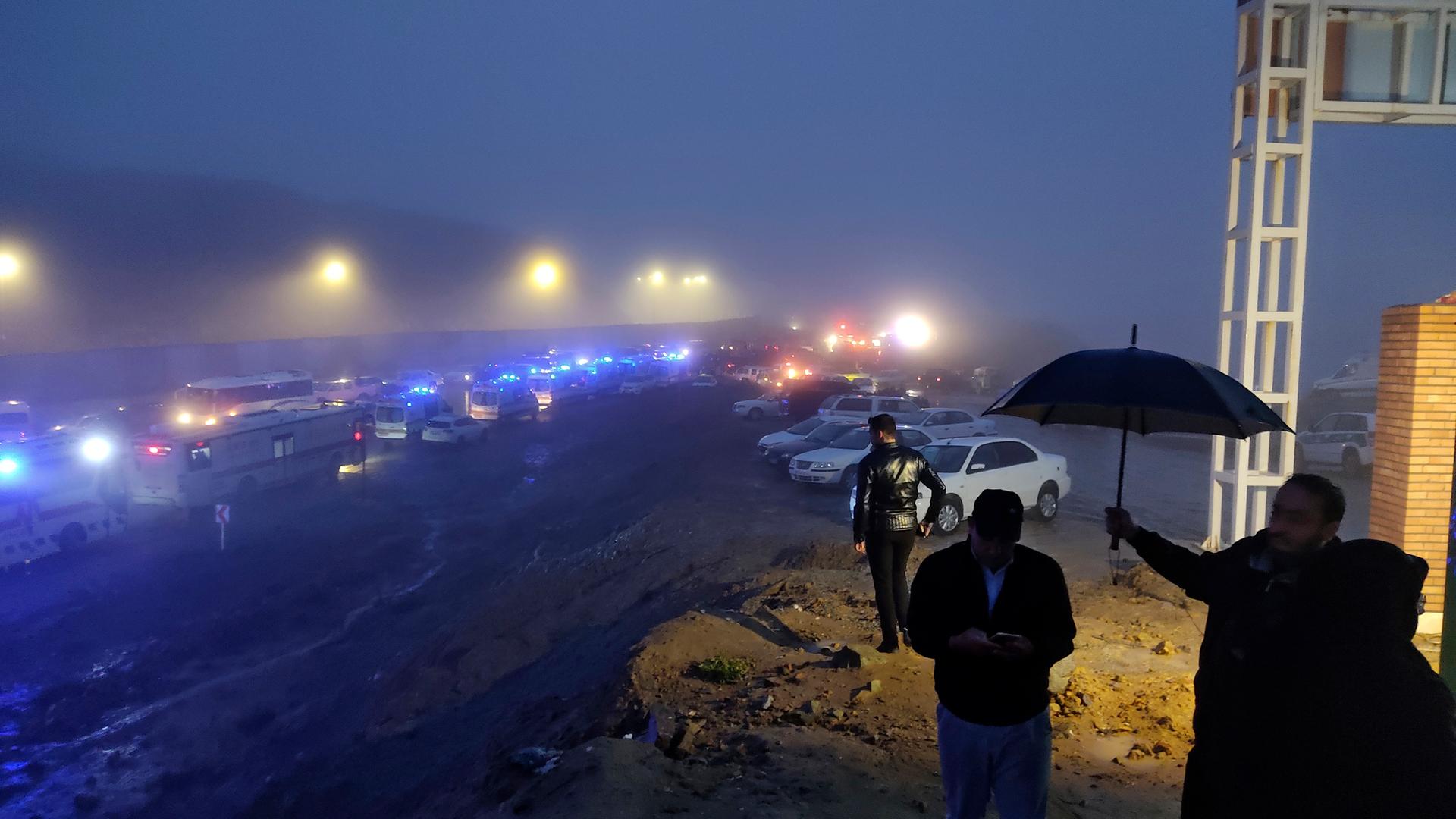 Einsatzfahrzeuge stehen nach dem Unglück mit einem Hubschrauber im Iran im Nebel. Im Vordergrund sind Menschen mit einem Regenschirm zu sehen. 