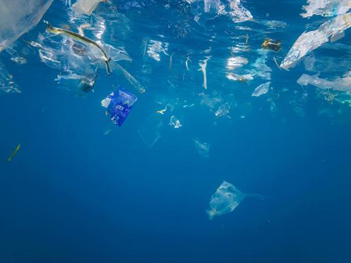 Plastiktüten, Plastikteile und Plastik schweben in strahlend blauem Wasser. Fische haben sich unter die Abfälle gemischt. Das Licht nimmt zum unteren Bildrand hin ab. 