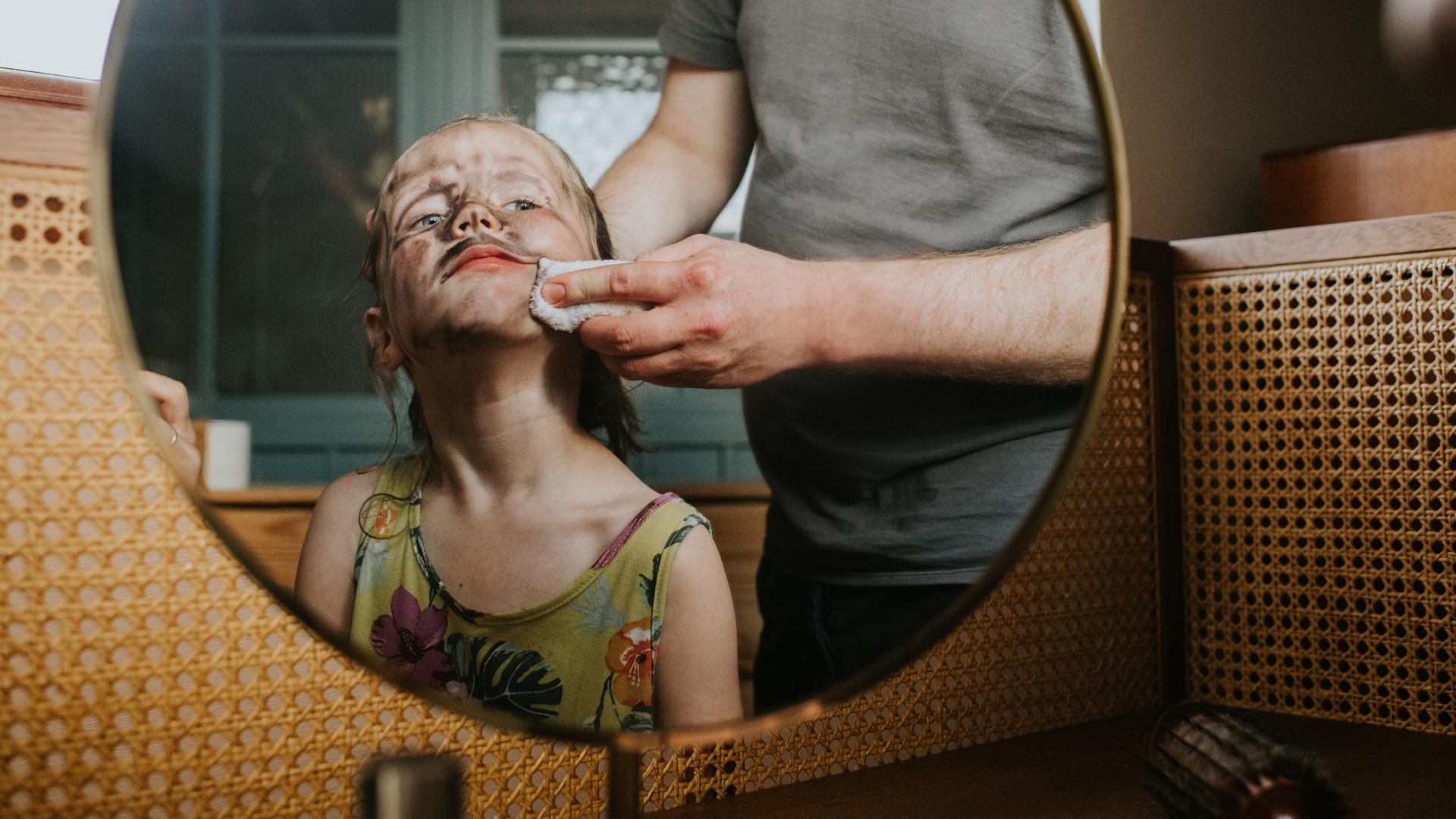 Blick in den Spiegel. Darin zu sehen ist ein Kind mit schmutzigem Gesicht und schelmischem Ausdruck. Eine Person schrubbt dem Kind mit einem Lappen das Gesicht.