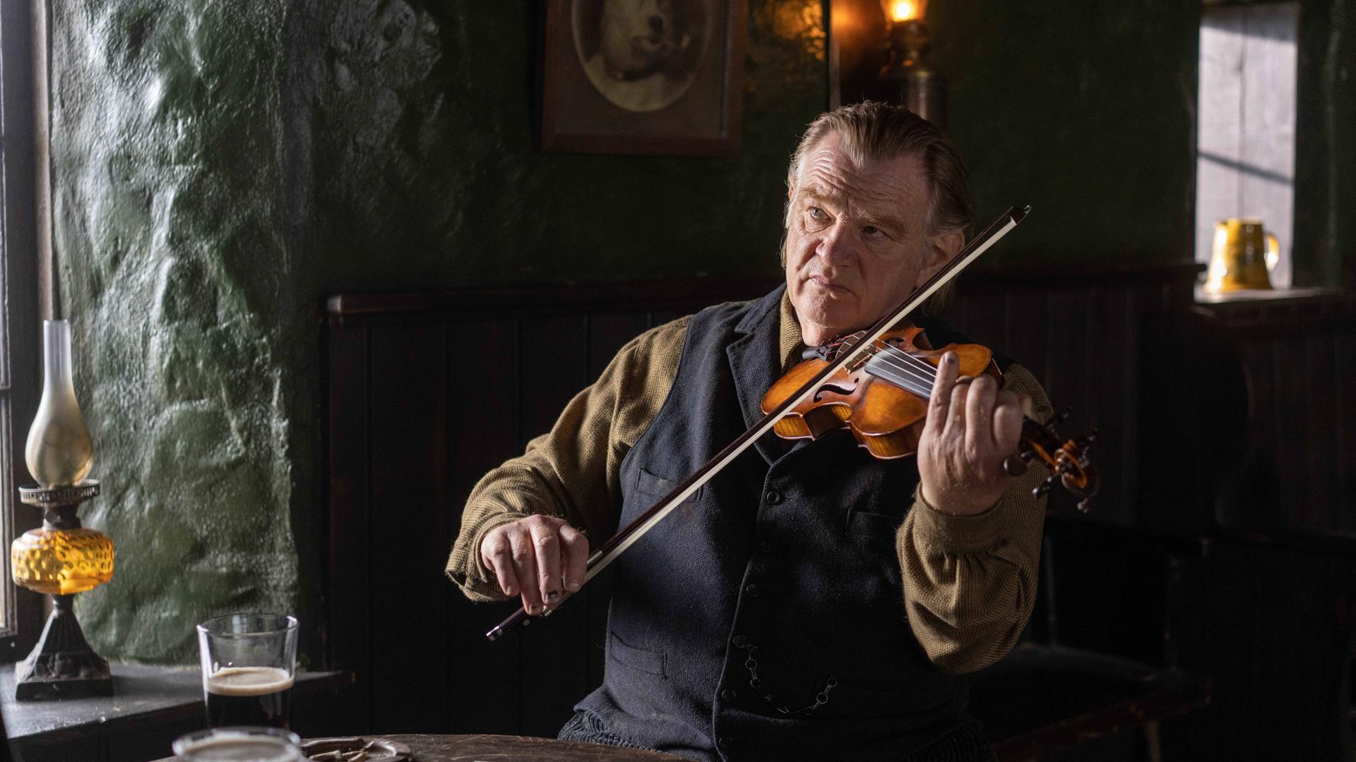 Szene aus dem Film "Banshees of Inisherin" mit dem irischen Schauspieler Brendan Gleeson, der in einem urigen Pub neben einem Fenster an einem Tisch vor einenm Glas Guinness sitzt und Geige spielt.