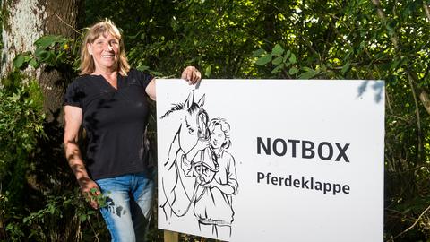 Pferdeklappe-Chefin Petra Teegen steht in Norderbrarup vor einem Schild mit der Aufschrift Notbox.
