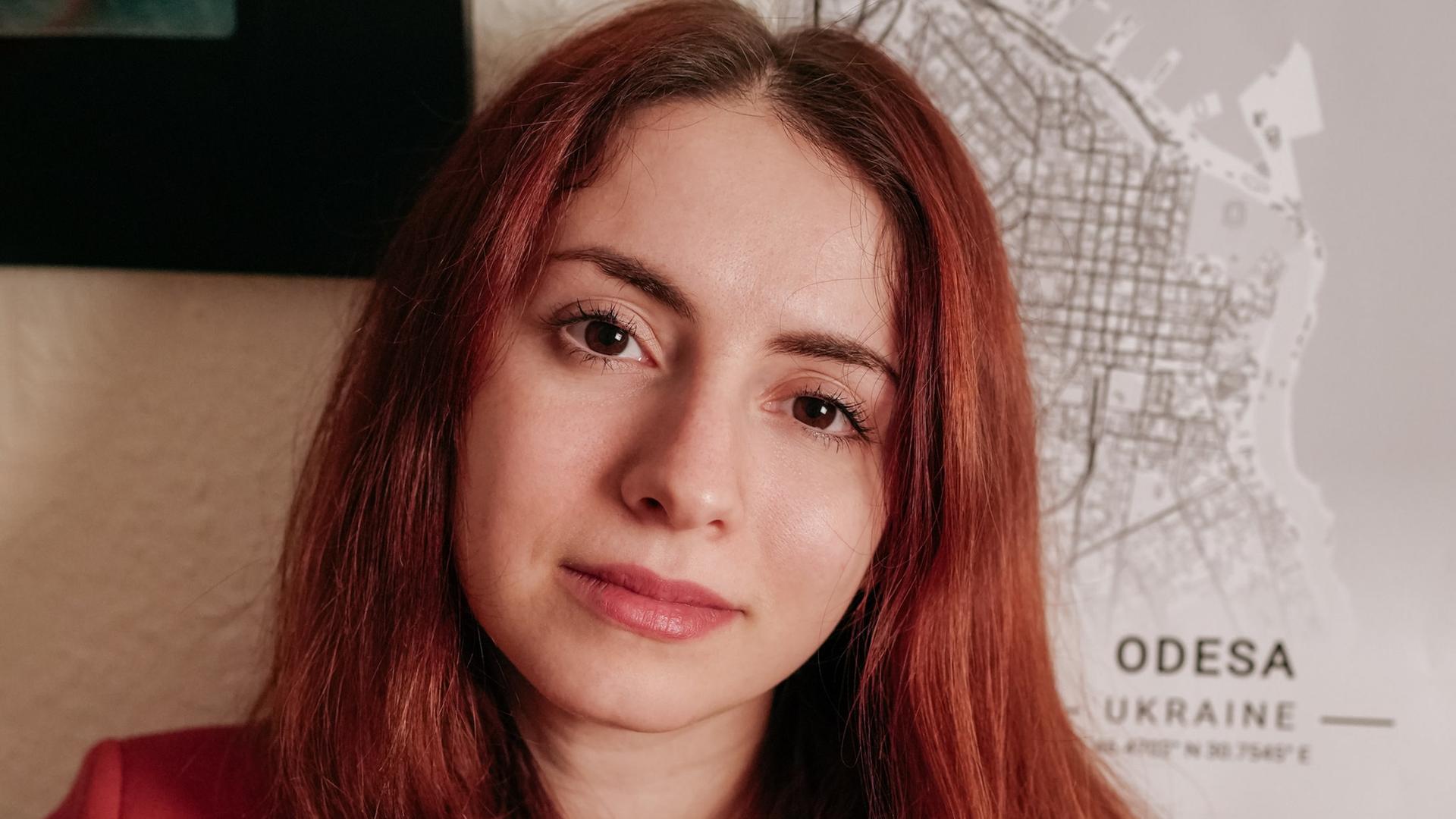 Die Ärztin und Autorin Iryna Fingerova steht in einem roten Pullover vor einer Wand, an der eine Karte von Odessa hängt.