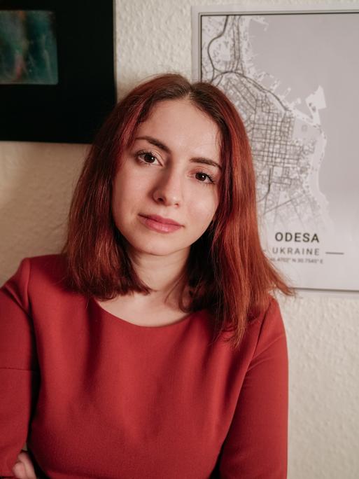 Die Ärztin und Autorin Iryna Fingerova steht in einem roten Pullover vor einer Wand, an der eine Karte von Odessa hängt.