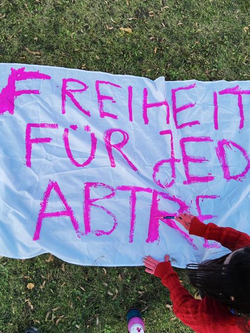 Eine Demonstrantin ist von oben zu sehen, die ein Bettlaken mit einem pinken Schriftzug bemalt, der bislang lautet: "Freiheit für jede Abtre..."