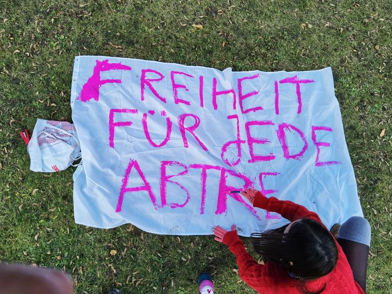 Eine Demonstrantin ist von oben zu sehen, die ein Bettlaken mit einem pinken Schriftzug bemalt, der bislang lautet: "Freiheit für jede Abtre..."
