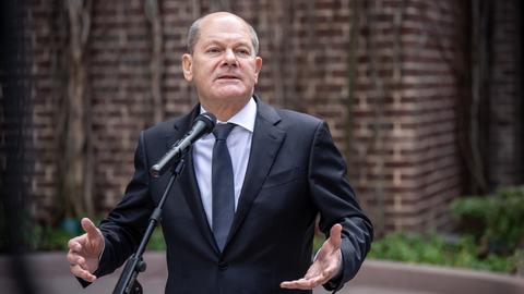 Scholz steht vor einem Mikrofon. Er trägt einen Anzug und eine schwarze Krawatte. Er gestikuliert mit beiden Händen.