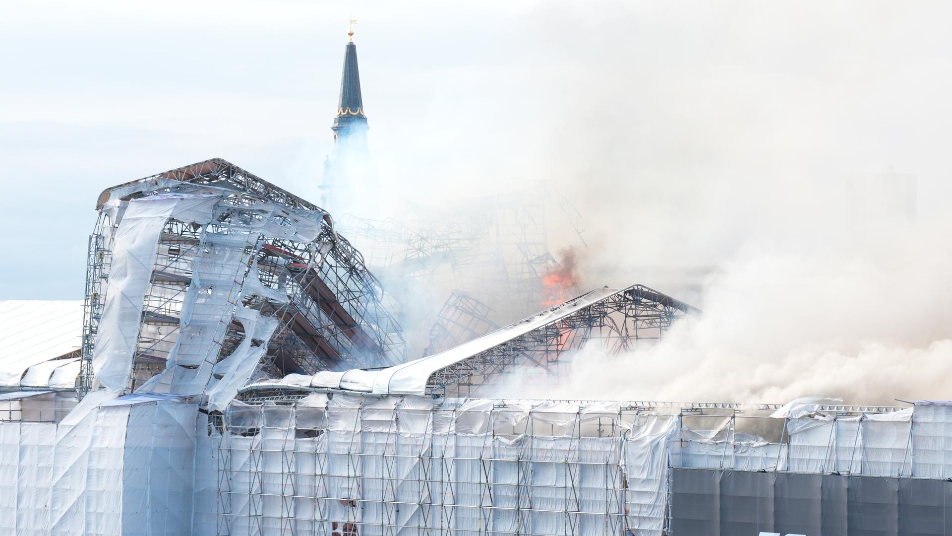 Dichter Rauch über der historischen Börse in Kopenhagen, die mit einem Baugerüst verhüllt ist. Flammen steigen aus der Mitte empor.