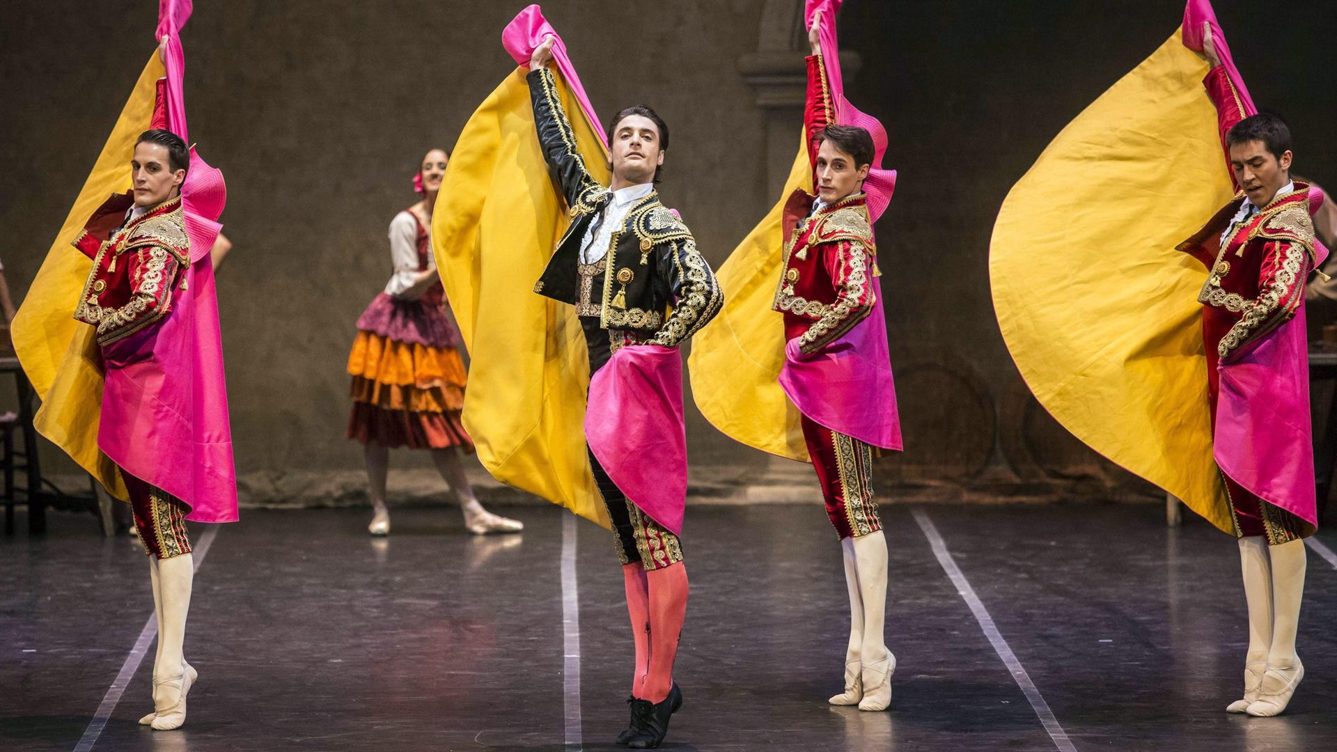 Vier Tänzer des Spanischen Staatsballetts stehen auf einer Bühne. Sie strecken sich von Kopf bis Fuß, stehen auf den Zehenspitzen und strecken ihre Arme in die Luft. Sie tragen verzierte Kleidung und pink-gelbe Umhänge.