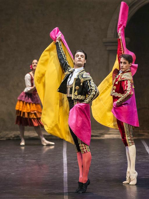 Vier Tänzer des Spanischen Staatsballetts stehen auf einer Bühne. Sie strecken sich von Kopf bis Fuß, stehen auf den Zehenspitzen und strecken ihre Arme in die Luft. Sie tragen verzierte Kleidung und pink-gelbe Umhänge.