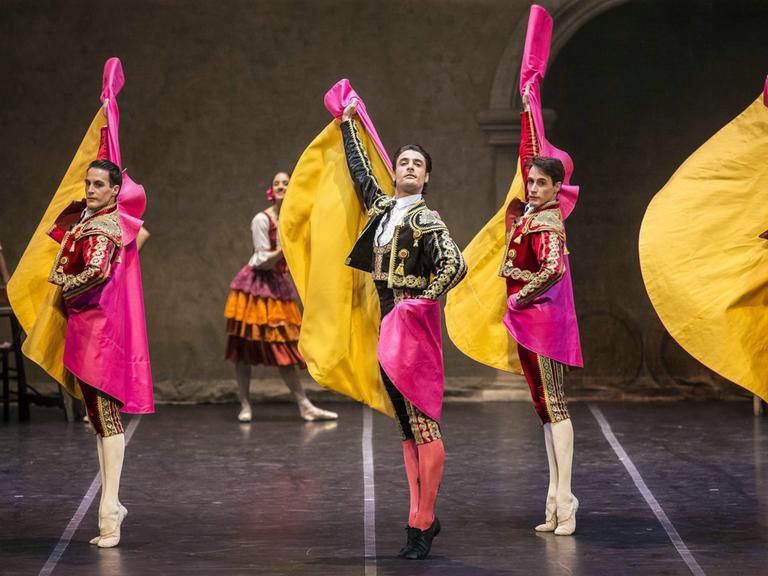 Vier Tänzer des Spanischen Staatsballetts stehen auf einer Bühne. Sie strecken sich von Kopf bis Fuß, stehen auf den Zehenspitzen und strecken ihre Arme in die Luft. Sie tragen verzierte Kleidung und pink-gelbe Umhänge.