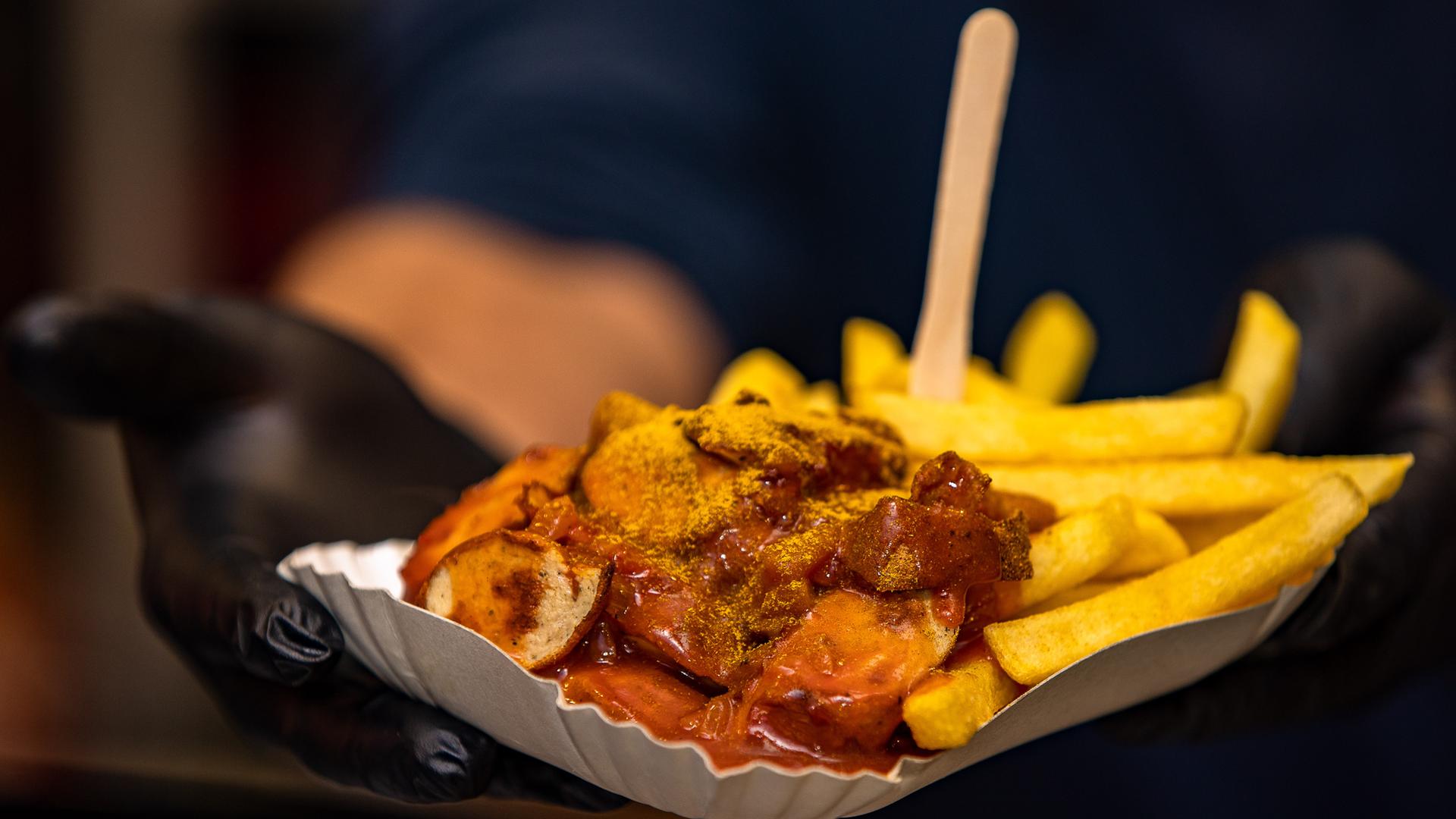 Currywurst und Pommes werden in einem Schälchen an einem Essensstand gerreicht.