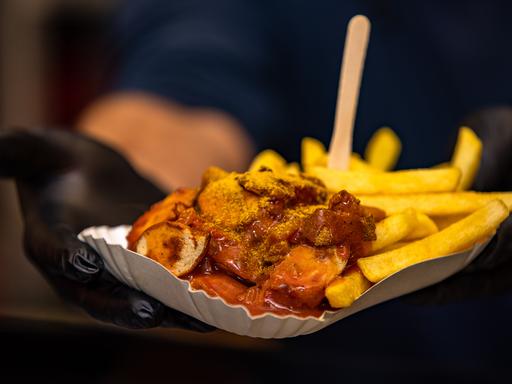 Currywurst und Pommes werden in einem Schälchen an einem Essensstand gerreicht.