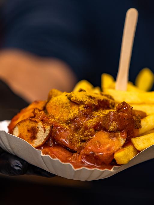 Currywurst und Pommes werden in einem Schälchen an einem Essensstand gerreicht.