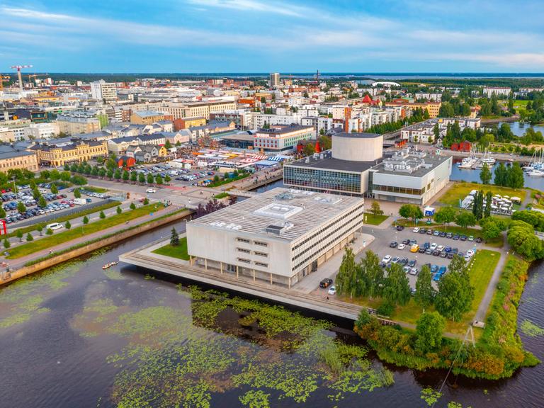 Panoramablick auf die finnische Stadt Oulu. Sie liegt am Wasser. Im Vordergrund sind große Gebäude aus Beton.