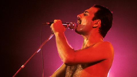Freddie Mercury steht mit freiem Oberkörper auf der Bühne, ist in rotes Licht getaucht und singt in ein Handmikro