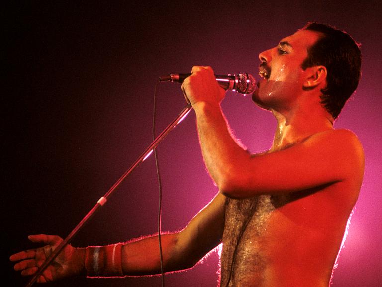 Freddie Mercury steht mit freiem Oberkörper auf der Bühne, ist in rotes Licht getaucht und singt in ein Handmikro Freddie Mercury steht mit freiem Oberkörper auf der Bühne, ist in rotes Licht getaucht und singt in ein Handmikro