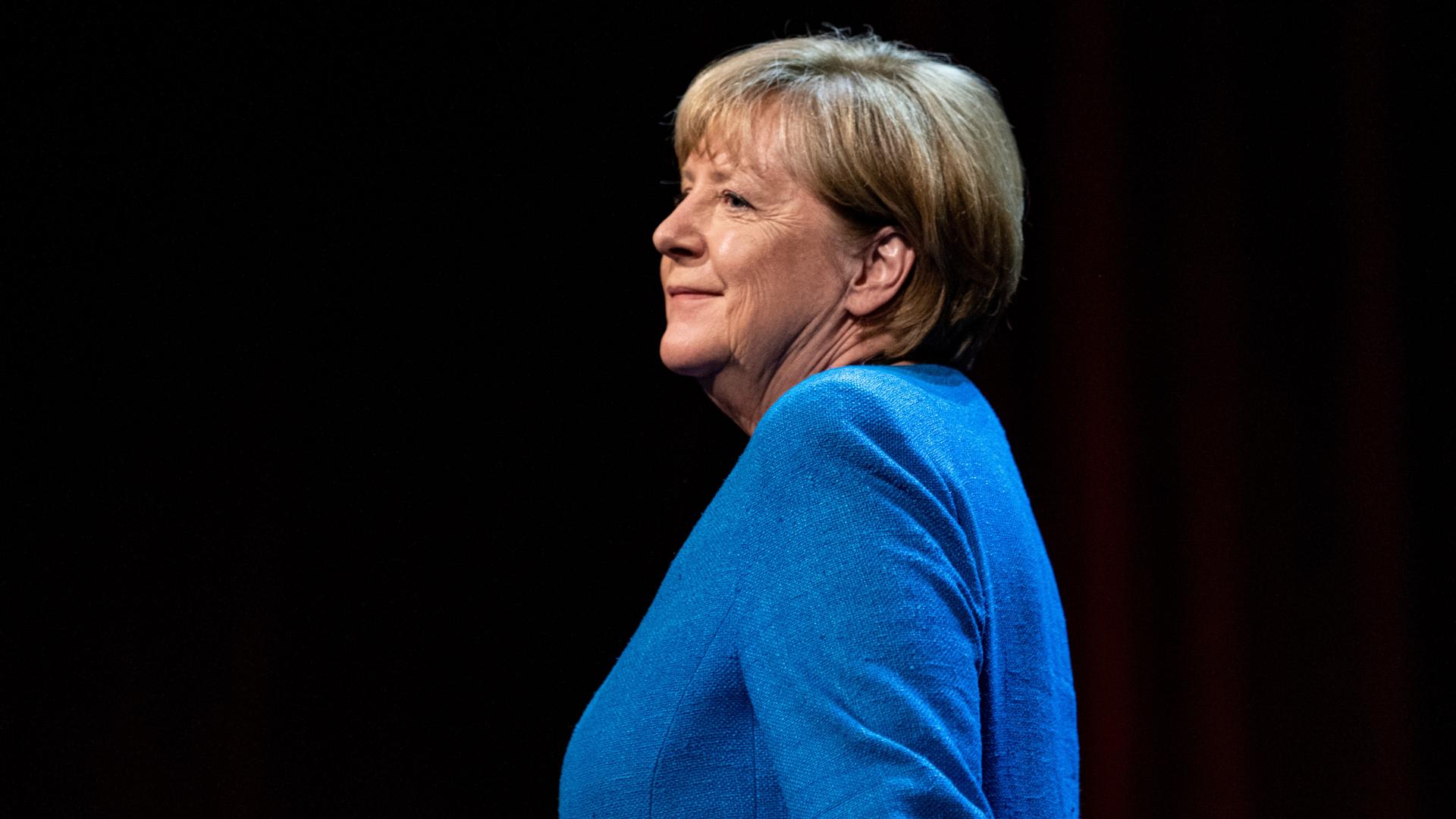 Seitliche Aufnahme der ehemaligen Bundeskanzlerin Angela Merkel (CDU) in einem blauen Blazer vor schwarzem Hintergrund.