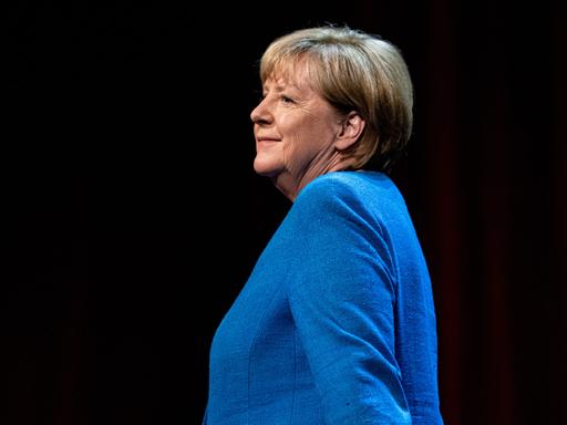 Seitliche Aufnahme der ehemaligen Bundeskanzlerin Angela Merkel (CDU) in einem blauen Blazer vor schwarzem Hintergrund.