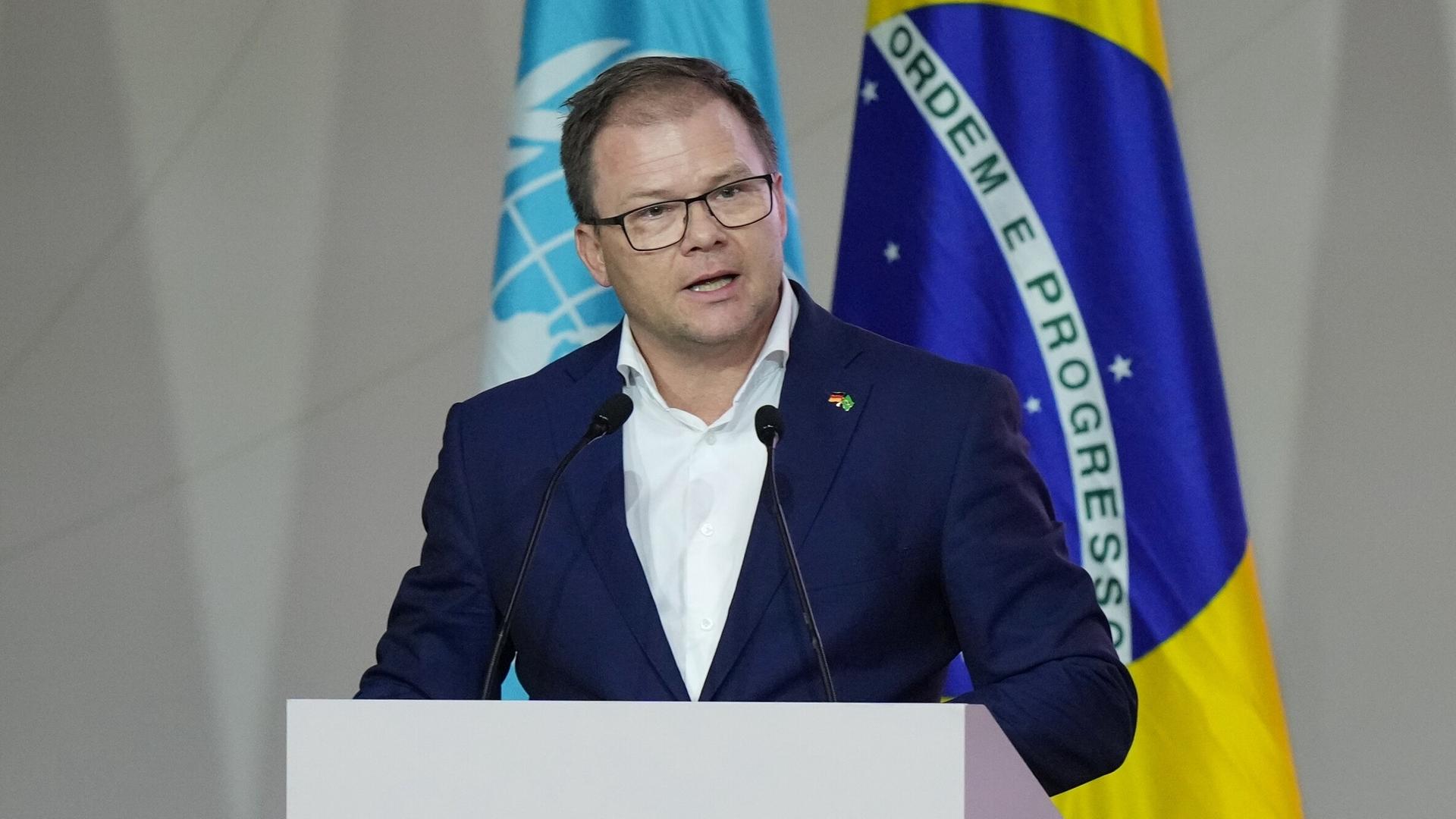 Carsten Schneider steht am Rednerpult, hinter ihm die Flaggen der UNO sowie Brasiliens.