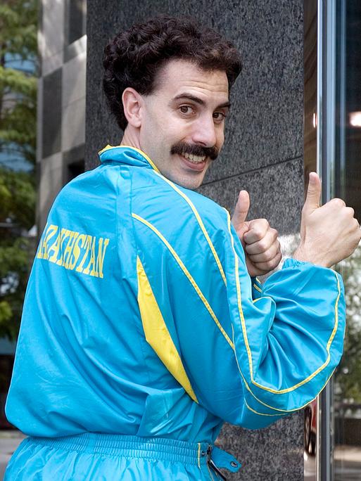 Der Schauspieler Sacha Baron Cohen als "Borat" in dem gleichnamigen Mockumentary-Film von 2006 schräg seitlich und teilweise von hinten zu sehen in einem blau-gelben Trainingsanzug auf dessen Rücken "Kasachstan" steht.