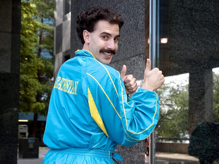 Der Schauspieler Sacha Baron Cohen als "Borat" in dem gleichnamigen Mockumentary-Film von 2006 schräg seitlich und teilweise von hinten zu sehen in einem blau-gelben Trainingsanzug auf dessen Rücken "Kasachstan" steht.