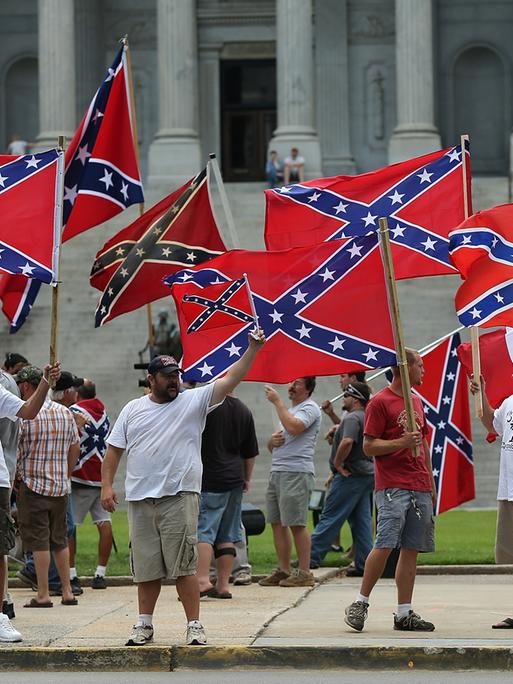 Demonstranten mit der Flagge der Konföderation (südstaaten Flagge) vor dem State House in South Carolina. Die Menschen stehen auf dem Bürgersteig und schwenken die Flaggen. (Symbolbild)