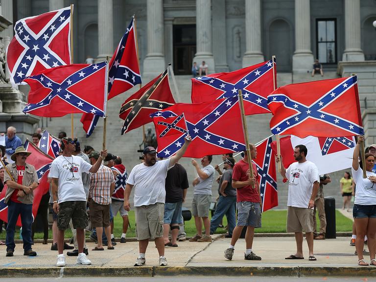 Demonstranten mit der Flagge der Konföderation (südstaaten Flagge) vor dem State House in South Carolina. Die Menschen stehen auf dem Bürgersteig und schwenken die Flaggen. (Symbolbild)