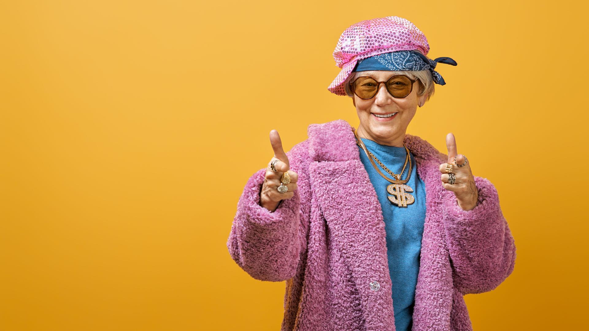 Eine Seniorin steht jugendlich gestylt vor einer knallgelben Wand. Sie trägt eine Kette mit glitzernder Dollarnote und einen pinken Pelzmantel.