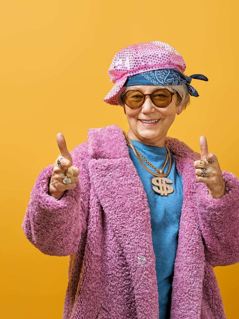 Eine Seniorin steht jugendlich gestylt vor einer knallgelben Wand. Sie trägt eine Kette mit glitzernder Dollarnote und einen pinken Pelzmantel.