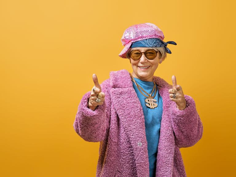 Eine Seniorin steht jugendlich gestylt vor einer knallgelben Wand. Sie trägt eine Kette mit glitzernder Dollarnote und einen pinken Pelzmantel.