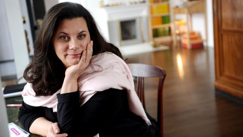 Tamara Dietl sitzt an einem Tisch, das Gesicht in die Hand gestützt. Sie hat schulterlanges dunkelbrauen Haar und trägt einen rosa Schal.