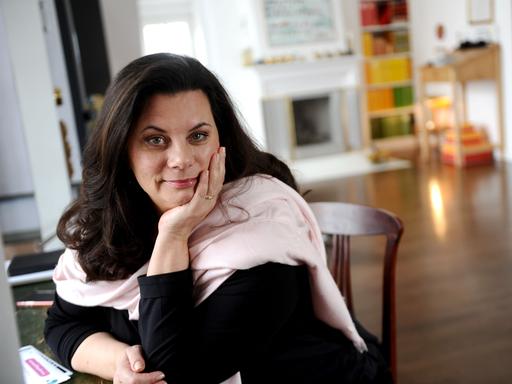 Tamara Dietl sitzt an einem Tisch, das Gesicht in die Hand gestützt. Sie hat schulterlanges dunkelbrauen Haar und trägt einen rosa Schal.