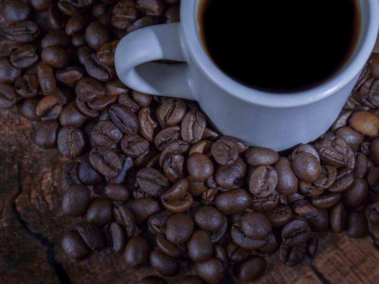 Weiße Keramiktasse mit dunklem Kaffee, die auf einem Bett aus Kaffeebohnen und einer rustikalen Holzoberfläche ruht