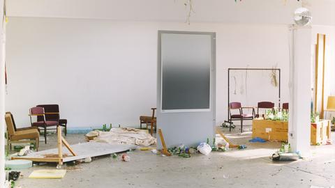 Eine Tür steht in der Mitte eines Zimmers, dass gerade renoviert wird. An der Decke hängt eine Discokugel. Überall stehen leere Flaschen.