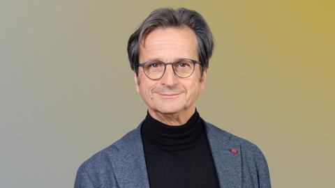 Ein Mann mit grauer, runder Brille und grauem Haar blickt lächelnd in die Kamera. Er trägt einen schwarzen Rollkragenpullover und ein graues Jackett.