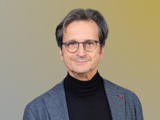 Ein Mann mit grauer, runder Brille und grauem Haar blickt lächelnd in die Kamera. Er trägt einen schwarzen Rollkragenpullover und ein graues Jackett.
