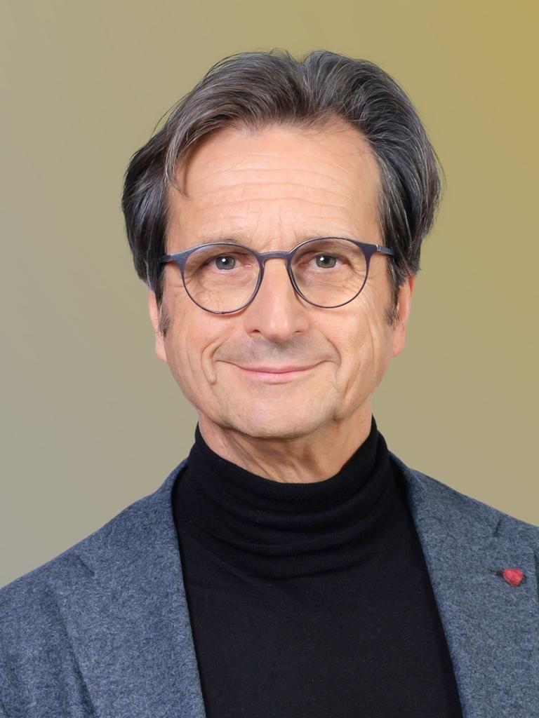 Ein Mann mit grauer, runder Brille und grauem Haar blickt lächelnd in die Kamera. Er trägt einen schwarzen Rollkragenpullover und ein graues Jackett.