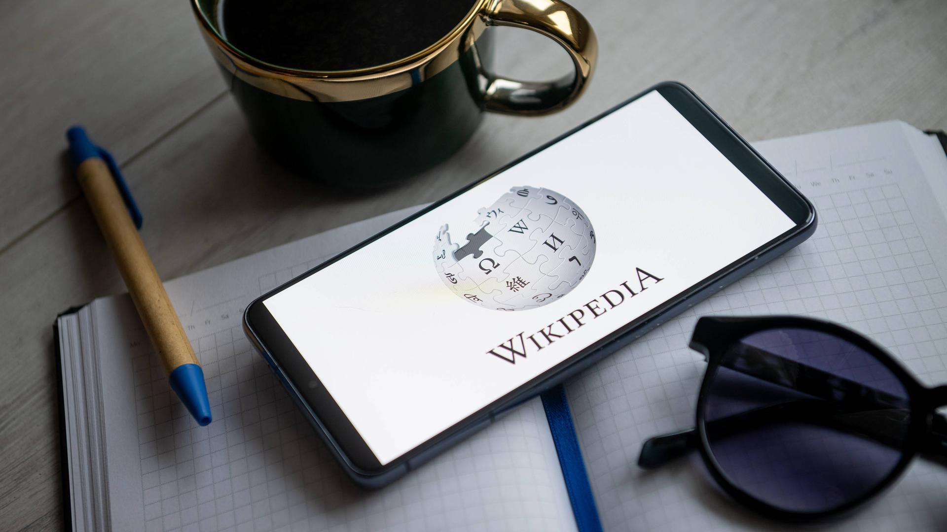 Ein Blick und eine Tasse und eine Sonnenbrille umrahmen ein Smartphone auf dessen Bildschirm das Wikipedia-Logo zu sehen ist