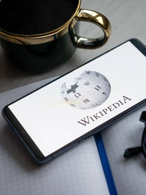 Ein Blick und eine Tasse und eine Sonnenbrille umrahmen ein Smartphone auf dessen Bildschirm das Wikipedia-Logo zu sehen ist