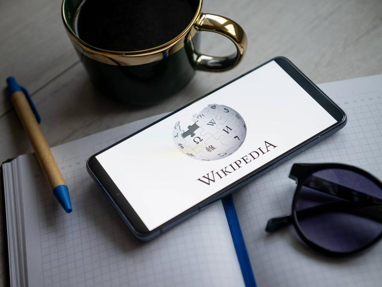 Ein Blick und eine Tasse und eine Sonnenbrille umrahmen ein Smartphone auf dessen Bildschirm das Wikipedia-Logo zu sehen ist