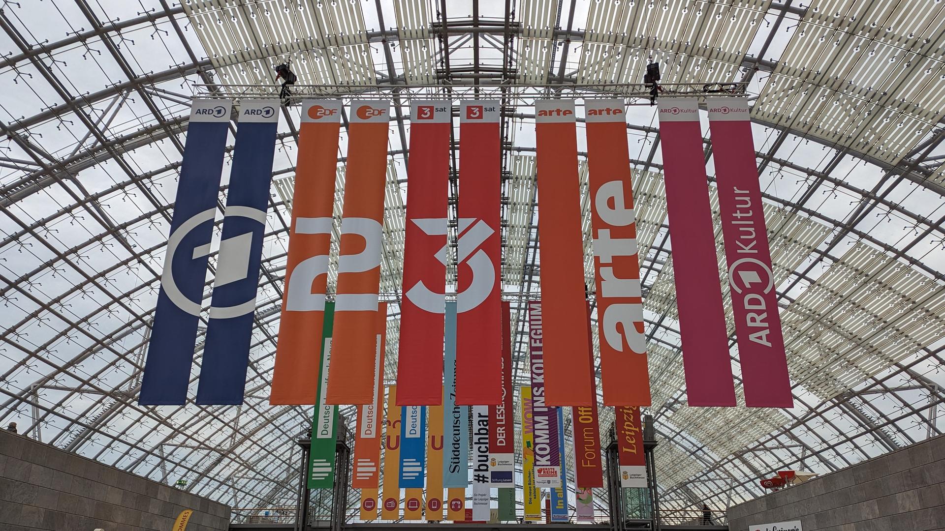 Bei der Leipziger Buchmesse hängen Banner der TV-Sender ARD, ZDF, 3Sat, Arte und ARD-Kultur von der Deke des Messegebäudes.