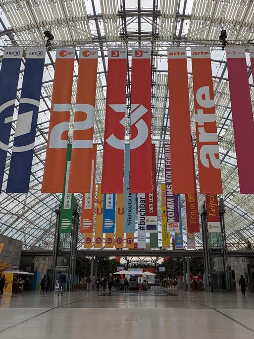 Bei der Leipziger Buchmesse hängen Banner der TV-Sender ARD, ZDF, 3Sat, Arte und ARD-Kultur von der Deke des Messegebäudes.